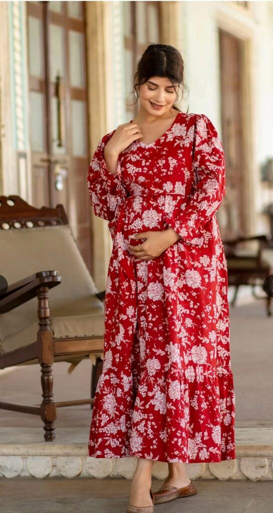 A pregnant woman looks radiant in a red floral printed maternity dress with long sleeves, posing gracefully while holding her baby bump.