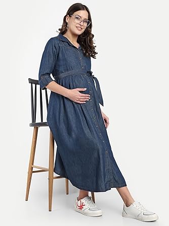 Pregnant woman wearing full-length denim maternity shirt dress with waist tie, button-down front, and white sneakers, sitting on wooden chair.